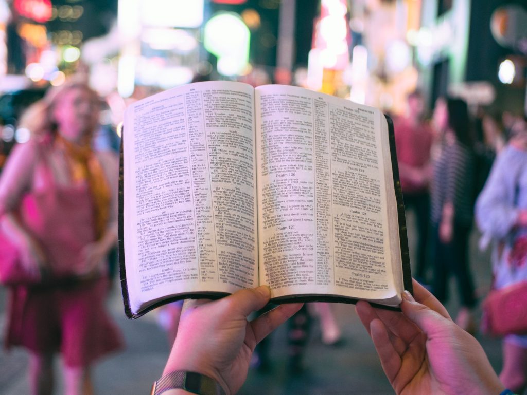 hope in difficult times: person holding bible on road with people walking on sidewalk beside buildings during nighttime