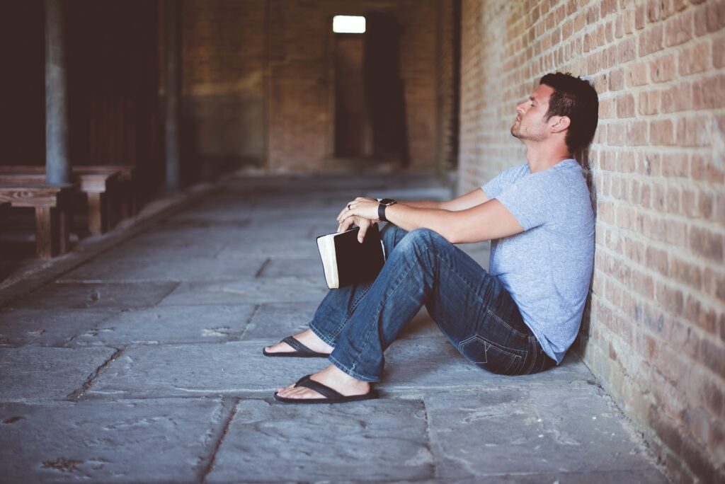 man sitting on pathway holding book at nighttime - Christian Wealth